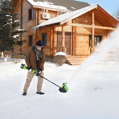Person using a snow blower in front of a wooden house with snow on the ground